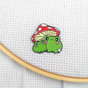 Aimant porte-aiguille illustré de deux grenouilles vertes sous un champignon rouge à pois, avec une étoile blanche